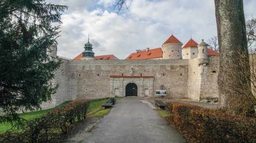 Zdjęcie przedstawia fasadę bramy z grubymi murami z majaczącym zamkiem ponad nimi.