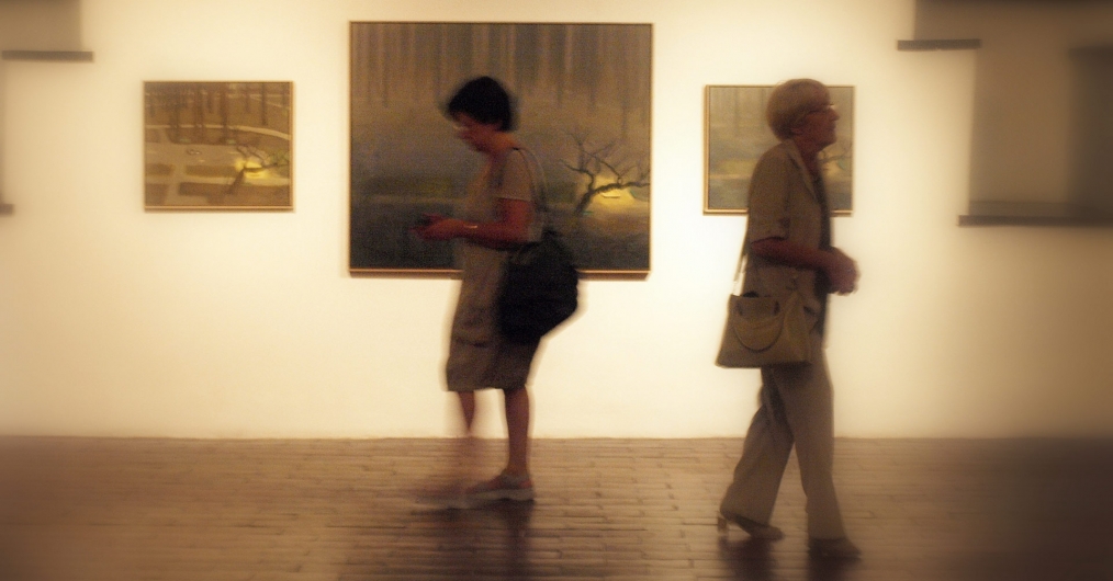 Zdjęcie lekko rozmazane, utrzymane w ciepłych tonach, przedstawia dwie kobiety idące korytarzem wystawy, wzdłuż ściany, na której wiszą zdjęcia w ramach.