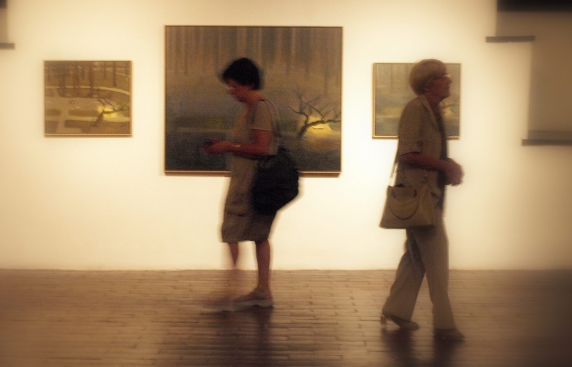 Zdjęcie lekko rozmazane, utrzymane w ciepłych tonach, przedstawia dwie kobiety idące korytarzem wystawy, wzdłuż ściany, na której wiszą zdjęcia w ramach.