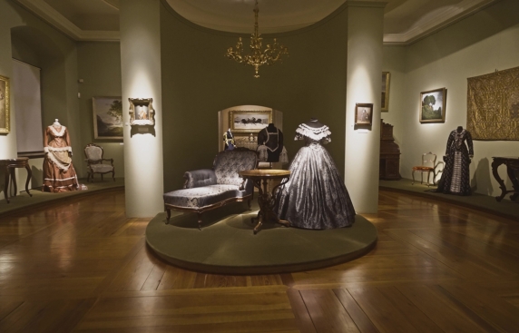 Zdjęcie przedstawia salę we wnętrzu zamku. Na środku ustawiony jest podest, na którym znajduje się szezlong, drewniany stolik i manekin z suknią z epoki.