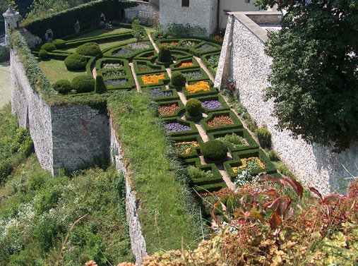 Zdjęcie przedstawia otoczony murami ogród w symetrycznej strukturze, znajdują się w nim rabaty z kolorowymi kwiatami.