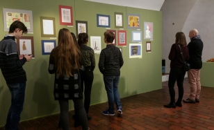 Zdjęcie przedstawia uczestników wystawy na tle ekspozycji. Na zielonej ścianie wywieszone są rysowane kredkami dziecięce obrazki.