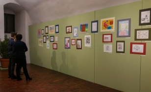 Zdjęcie przedstawia uczestników wystawy na tle ekspozycji. Na zielonej ścianie wywieszone są rysowane kredkami dziecięce obrazki w kolorowych ramkach.