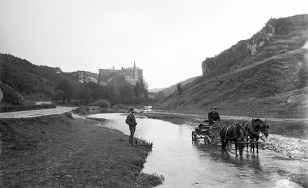 Czarno-biała fotografia przedstawiająca strumień i stojącego na jego brzegu mężczyznę oraz wóz zaprzęgnięty dwoma końmi i powożącego nim mężczyznę. W tle skalne zbocza oraz zamek.