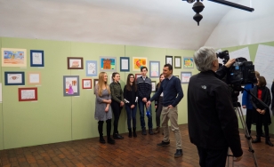 Zdjęcie przedstawia uczestników wystawy na tle ekspozycji. Na zielonej ścianie wywieszone są rysowane kredkami dziecięce obrazki w kolorowych ramkach.