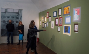 Zdjęcie przedstawia uczestników wystawy na tle ekspozycji. Na zielonej ścianie wywieszone są rysowane kredkami dziecięce obrazki.