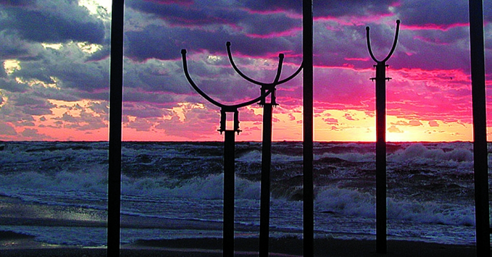 Zdjęcie przedstawia morską panoramę o zachodzie słońca. Morskie fale rozbijają się o brzeg, a na pierwszym planie widnieje instalacja składająca się z drewnianych słupów. Niebo przybiera kolory pomarańczu i różu, na nim niebieskie chmury.