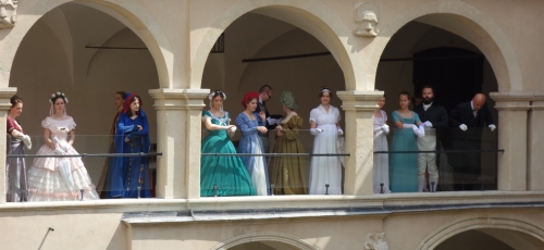 Zdjęcie przedstawia uczestników wydarzenia ubranych w stroje z epoki, stojących na krużgankach.