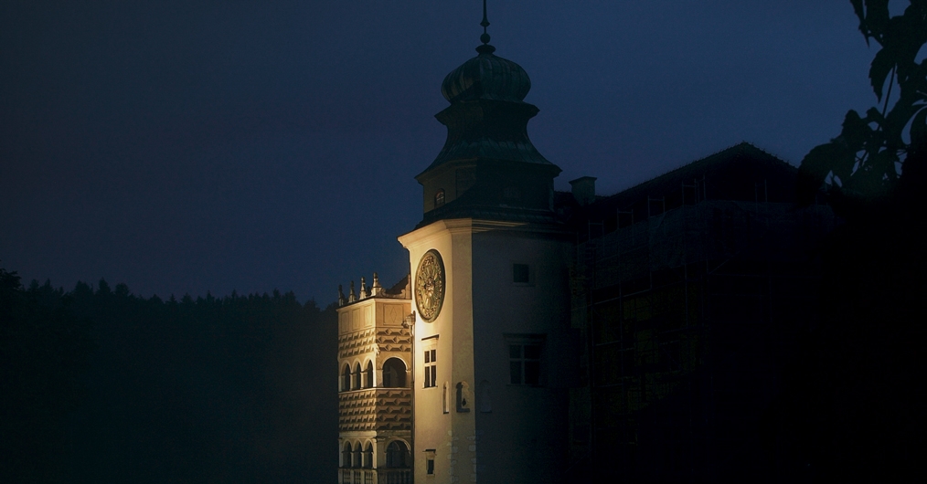 Zdjęcie wydarzenia Noc w muzeum, przedstawia zamek nocą z oświetloną wieżą zegarową i krużgankami.