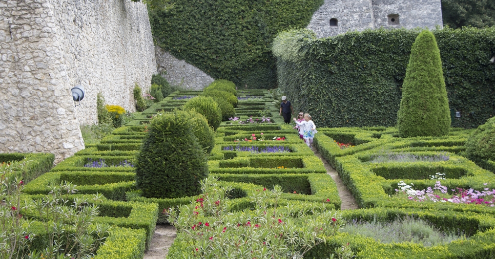 Zdjęcie przedstawia zielony ogród złożony z symetrycznych struktur z bukszpanu, przypominających labirynt. W ogrodzie znajdują się także przycięte krzaki oraz klomby kwiatów.