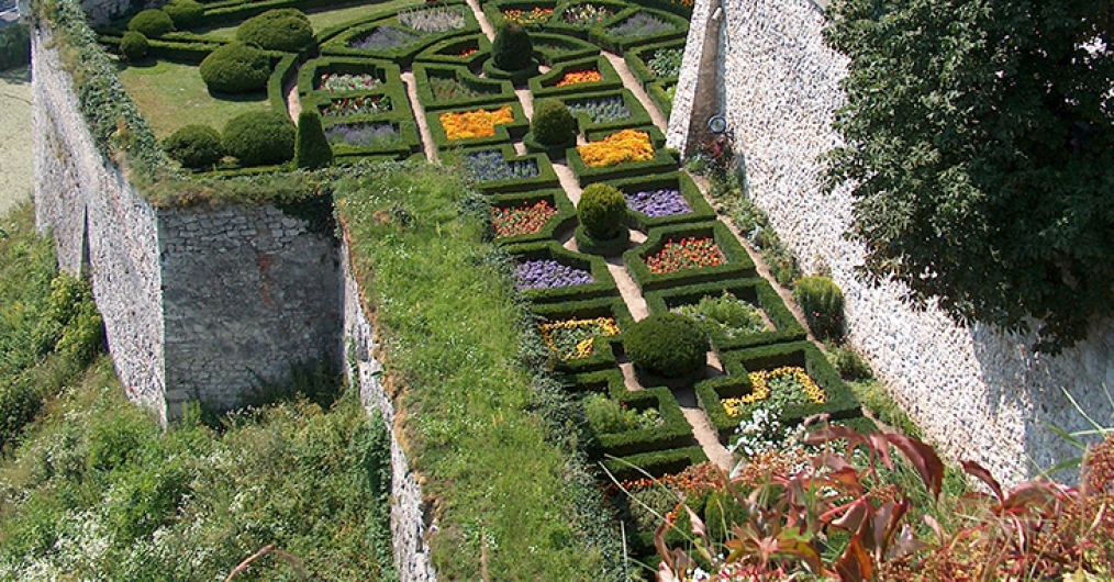 Zdjęcie przedstawia zielony ogród złożony z symetrycznych struktur z bukszpanu, przypominających labirynt. W ogrodzie znajdują się także przycięte krzaki oraz klomby kwiatów.