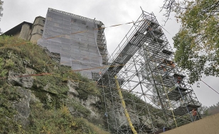 Zdjęcie przedstawia zamek widziany od dołu podczas renowacji. Na pierwszym planie porośnięta mchem i krzewami skała oraz rusztowanie.