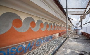 Zdjęcie przedstawia malowidło na murze podczas renowacji. Malowidło składa się z dwóch szerokich pasów - żółtego i pomarańczowego, na ich złączeniu widnieją szare geometryczne wzory przypominające fale. Obok stoi rusztowanie.