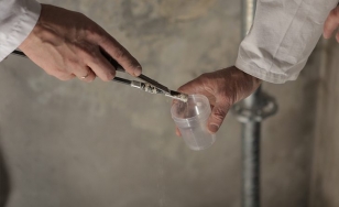 Zdjęcie przedstawia dwie dłonie umieszczające próbkę tynku w plastikowym, cylindrycznym opakowaniu.