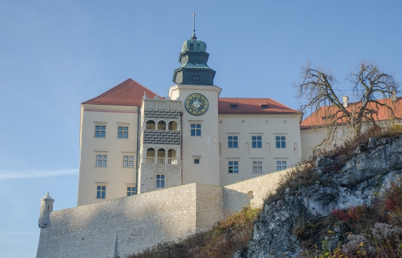Zdjęcie przedstawia ścianę zamku z perspektywy podnóżna wzgórze, widoczna jest wieża zegarowa i zdobiona loggia z arkadami.