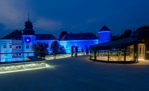 Zdjęcie przedstawia zamek w Pieskowej Skale wieczorową porą. Zamek oświetlony jest niebieskim światłem, a stojąca na pierwszym planie altanka podświetlona jest na żółto.