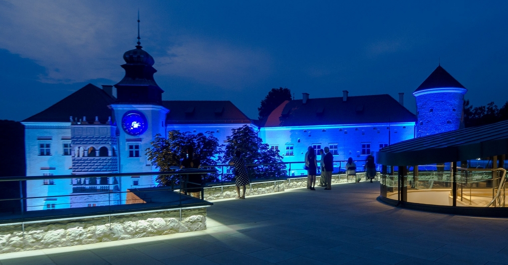 Zdjęcie przedstawia zamek w Pieskowej Skale wieczorową porą. Zamek oświetlony jest niebieskim światłem, a stojąca na pierwszym planie altanka podświetlona jest na żółto.