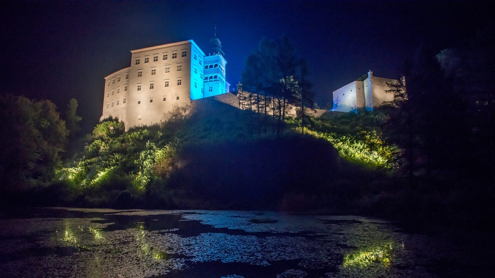 Zdjęcie przedstawia nocny zamek z żabiej perspektywy, oświetlony jest białym i niebieskim światłem, tworząc łunę wokół wieży zamkowej.