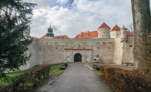 Zdjęcie przedstawia bramę prowadzącą do Zamku w Pieskowej Skale. Boki dróżki dojazdowej porastają krzewy. Zza muru wyłaniają się czerwone dachy zamku.