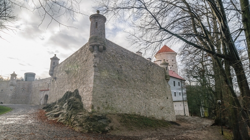 Zdjęcie przedstawia róg muru zamku z niewielkimi wieżyczkami, niebo jest pochmurne, a w otoczeniu znajdują się bezlistne drzewa.