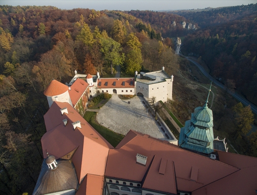 Zdjęcie przedstawia zamek z lotu ptaka, widoczny jest brukowany dziedziniec oraz dachówki w ceglastym kolorze. W tle znajduje się rozległy, jesienny las.