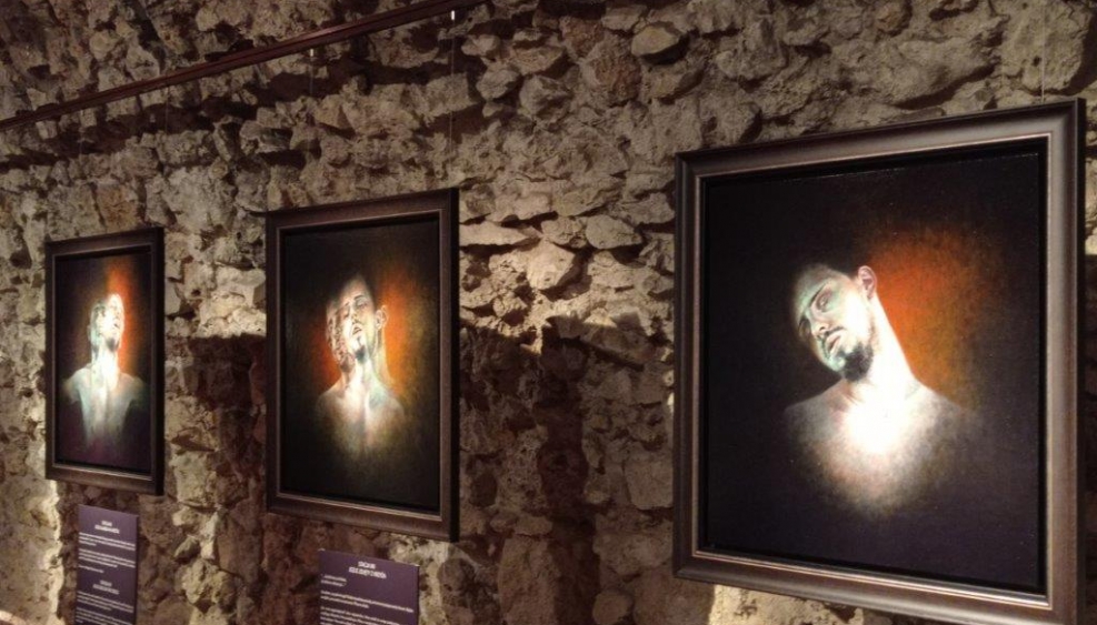 sala wystawy oświetlona nastrojowym światłem, ściana z kamienia, na niej zawieszone trzy obrazy z portretem mężczyzny