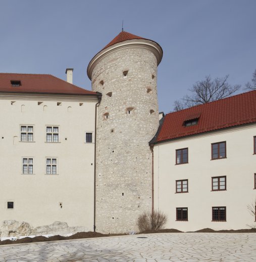 Na zdjęciu widoczna jest prosta wieża z dachem w kolorze ceglastym, znajduje się pomiędzy dwoma budynkami.