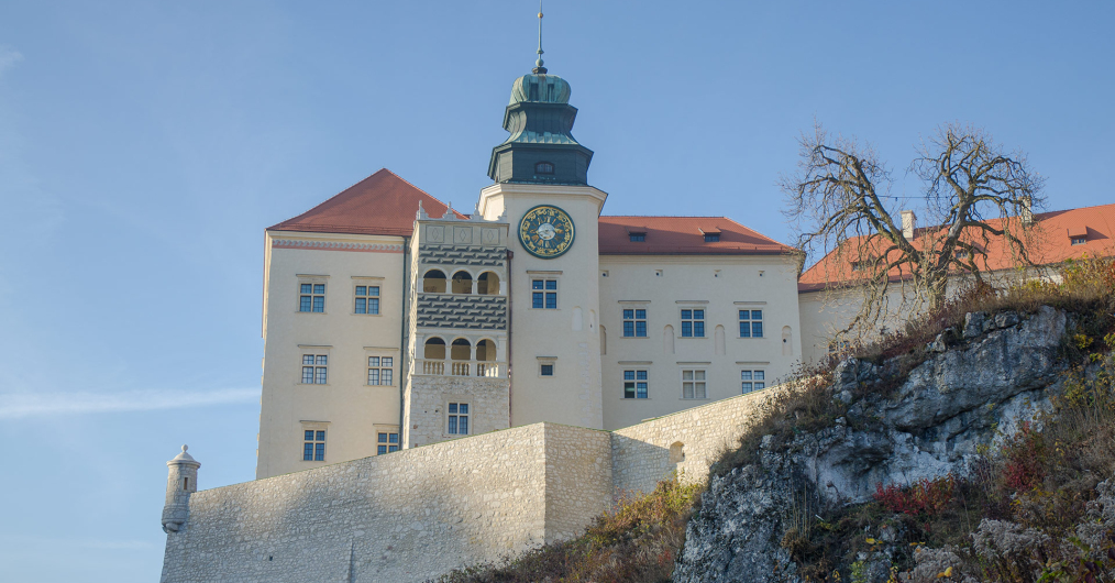 Zdjęcie przedstawia ścianę zamku z perspektywy podnóżna wzgórze, widoczna jest wieża zegarowa i zdobiona loggia z arkadami.