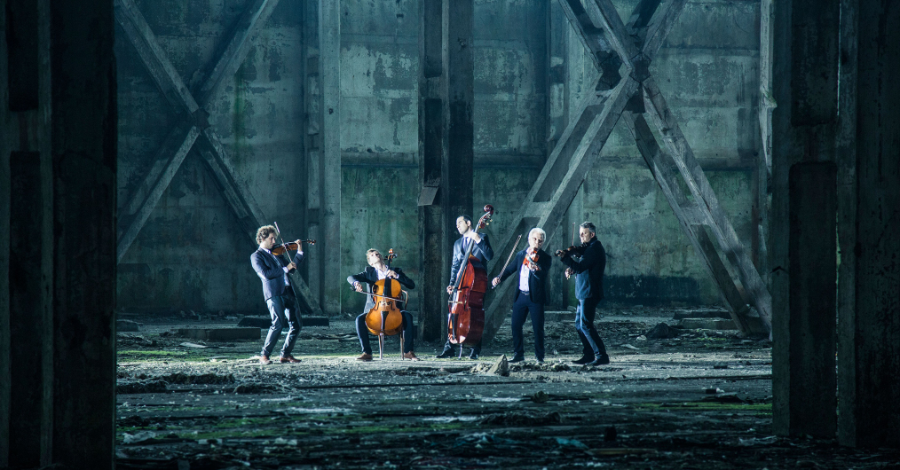 grupa muzyków grających na różnych instrumentach, w przestrzeni jakiegoś magazynu lub hali fabrycznej