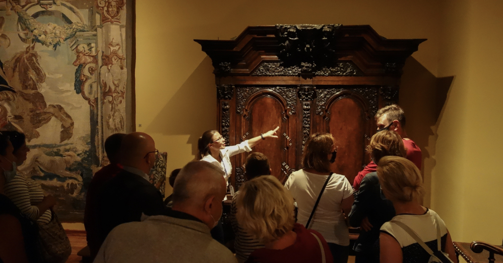 Zwiedzający muzeum przyglądają się zabytkowej szafie i tkaninie na ścianie