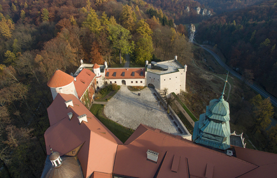 Zdjęcie przedstawia zamek z lotu ptaka, widoczny jest brukowany dziedziniec oraz dachówki w ceglastym kolorze. W tle znajduje się rozległy, jesienny las.
