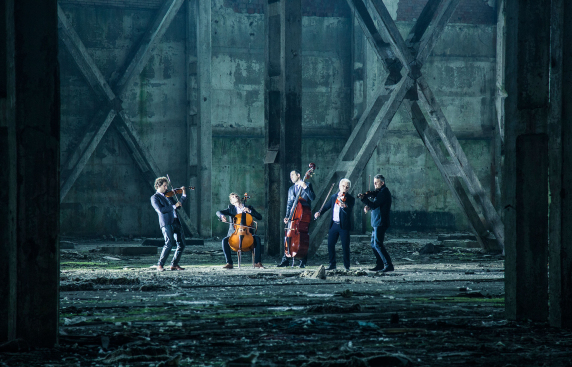 grupa muzyków grających na różnych instrumentach, w przestrzeni jakiegoś magazynu lub hali fabrycznej