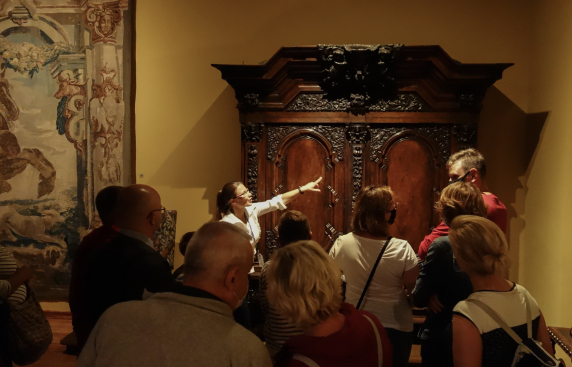 Zwiedzający muzeum przyglądają się zabytkowej szafie i tkaninie na ścianie